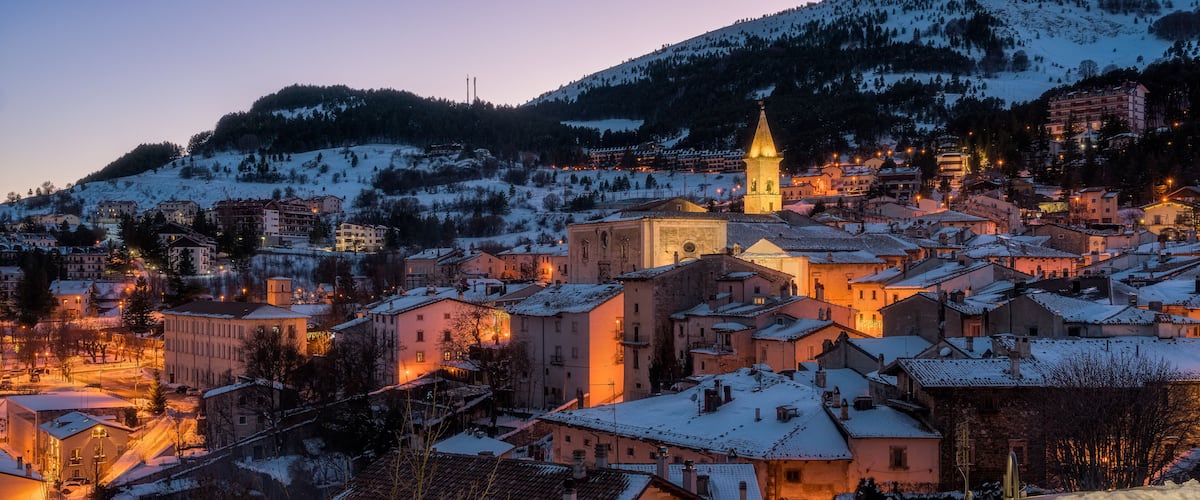 A snow-covered town at dusk with illuminated buildings and a prominent church tower.