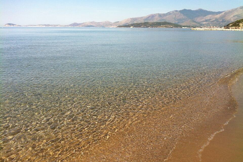 A clear beach with shallow water, a sandy shore, and distant mountains.