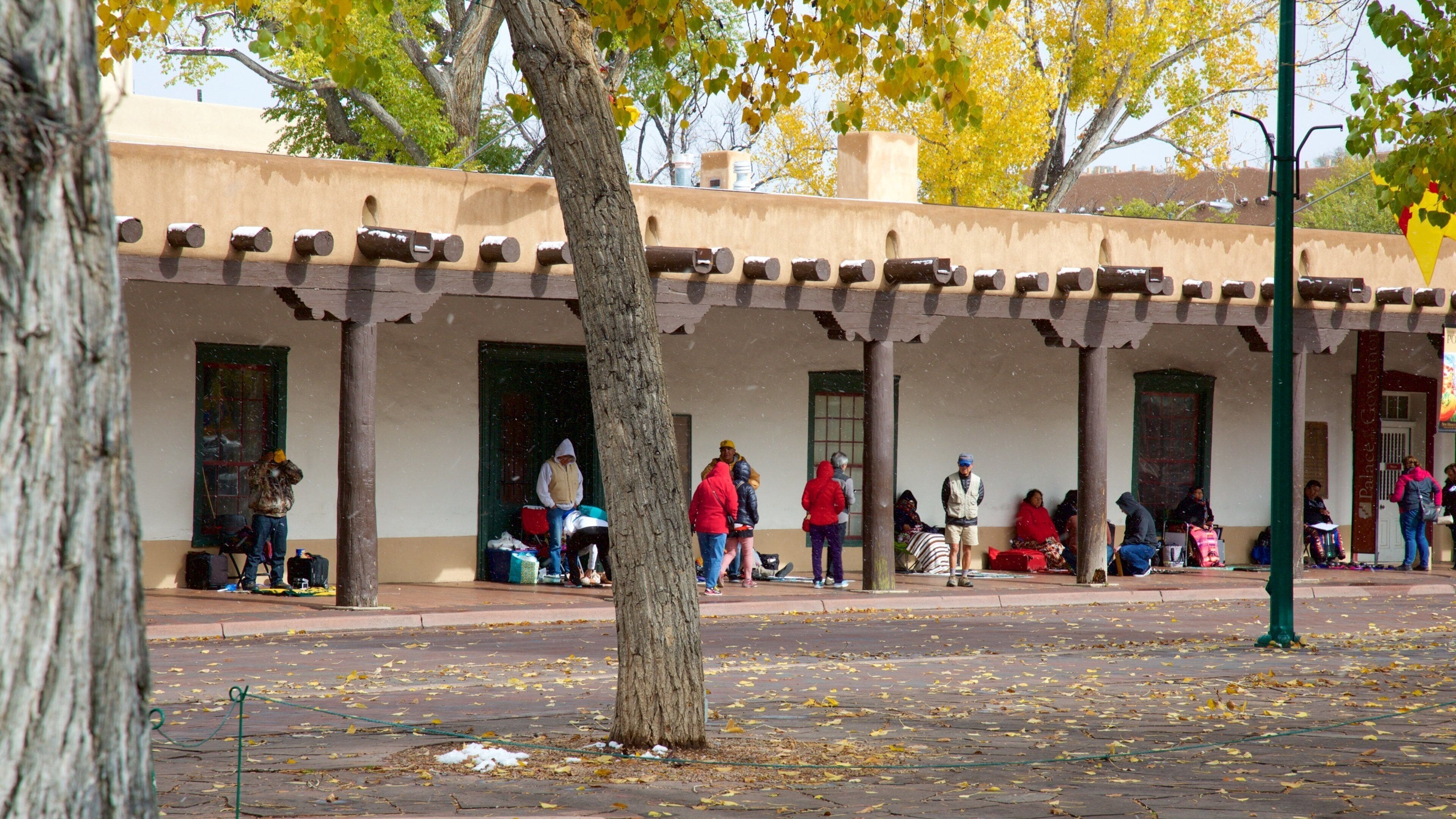 A plaza with a covered building, trees with yellow leaves, and people sitting and standing.
