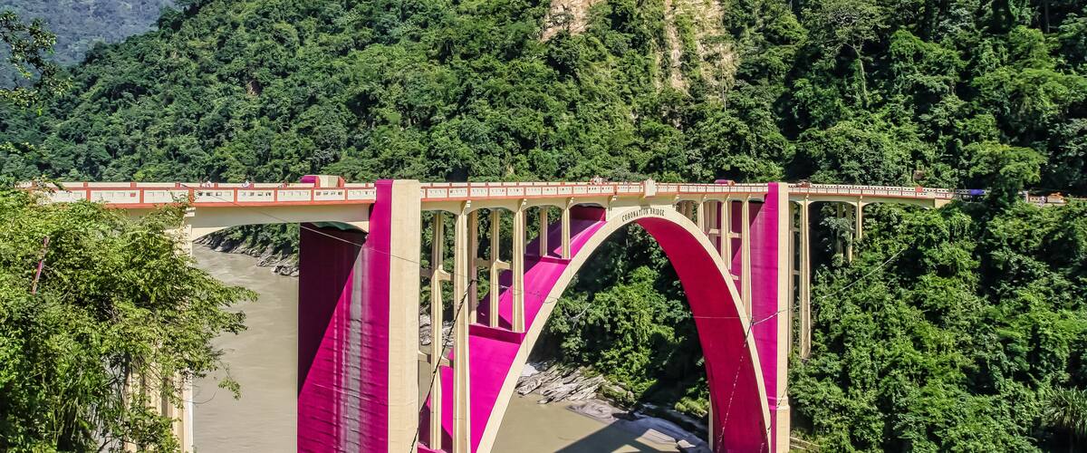 A bridge with pink arches over a river, surrounded by lush green hills.