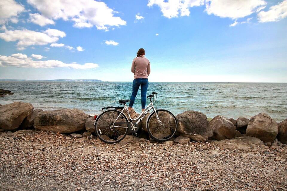 A woman standing by a bicycle on a rocky shore with the ocean in the background.