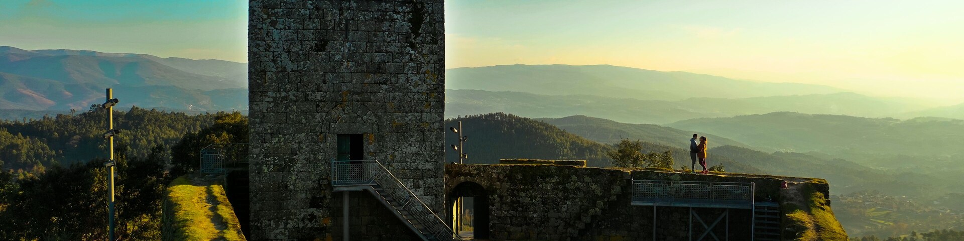 Una torre de piedra con una escalera, rodeada de césped y vistas a montañas distantes.