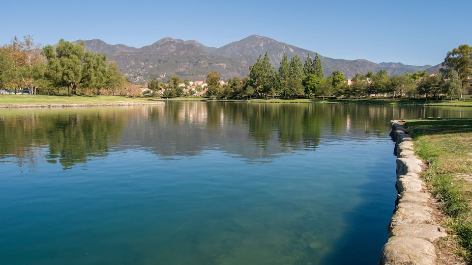 A calm lake with clear reflections, surrounded by trees and mountains in the background.