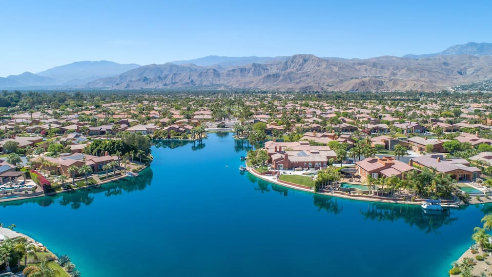 A residential area with a large body of water, surrounded by houses and palm trees, with mountains in the background.