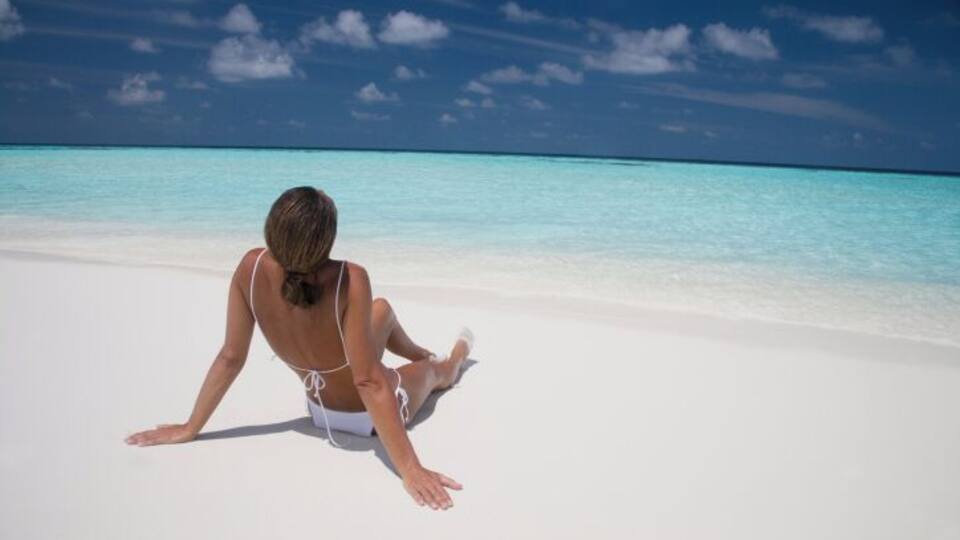 A person sitting on a white sandy beach with clear turquoise water and a blue sky.