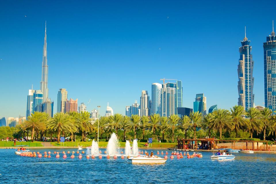 A city skyline with modern skyscrapers, a body of water with boats and fountains, and palm trees along the shore.