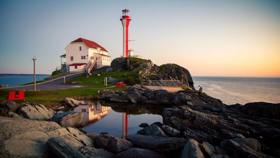 A lighthouse on a rocky outcrop with a white building nearby, a calm sea, and a clear sky.