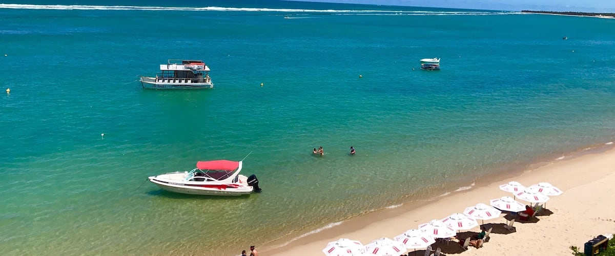 A beach with clear turquoise water, a boat anchored near the shore, and beach umbrellas lined up along the sand.