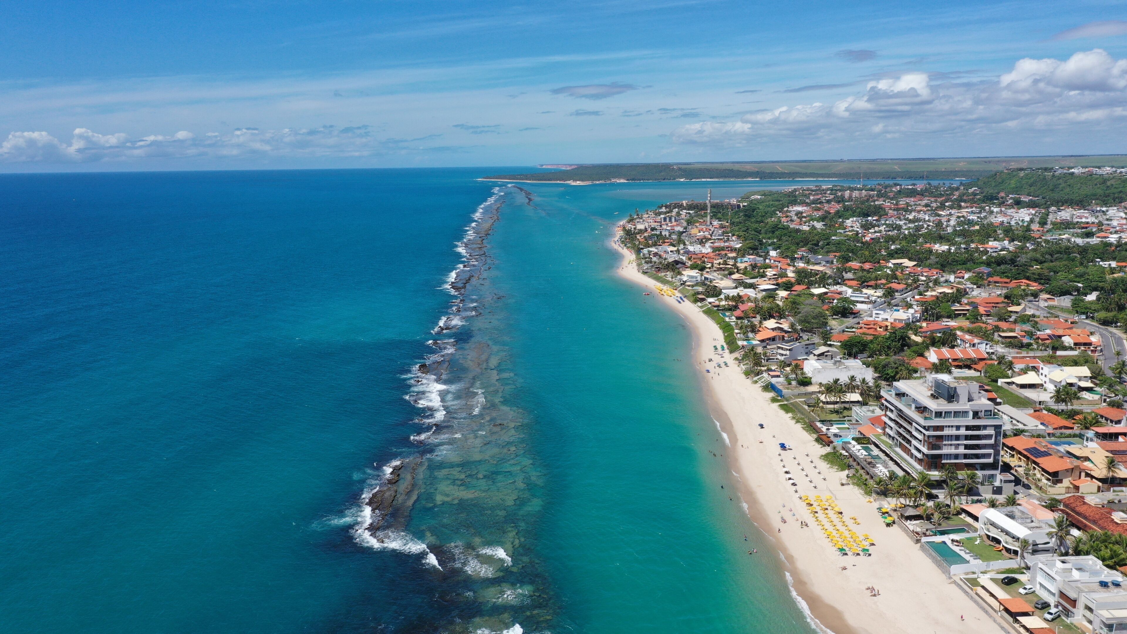 A coastal town with a long beach, clear blue waters, and a dense residential area with colorful buildings.