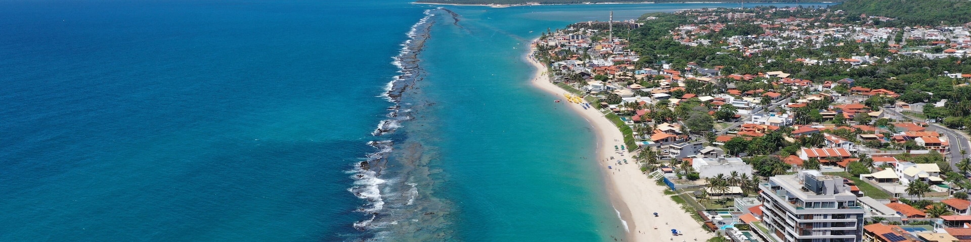 A coastal town with a long beach, clear blue waters, and a dense residential area with colorful buildings.