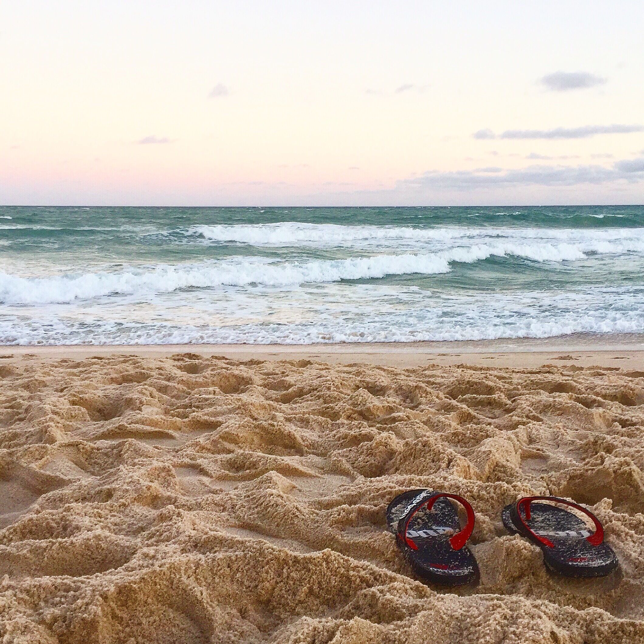 A beach with waves and a pair of flip-flops on the sand.