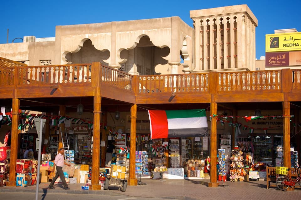 Un mercado tradicional con balcones de madera, tiendas que ofrecen diversos productos y una bandera de los Emiratos Árabes Unidos.
