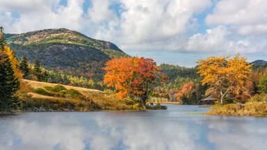 A lake surrounded by autumn-colored trees, a hill in the background, and a small cabin near the water's edge.