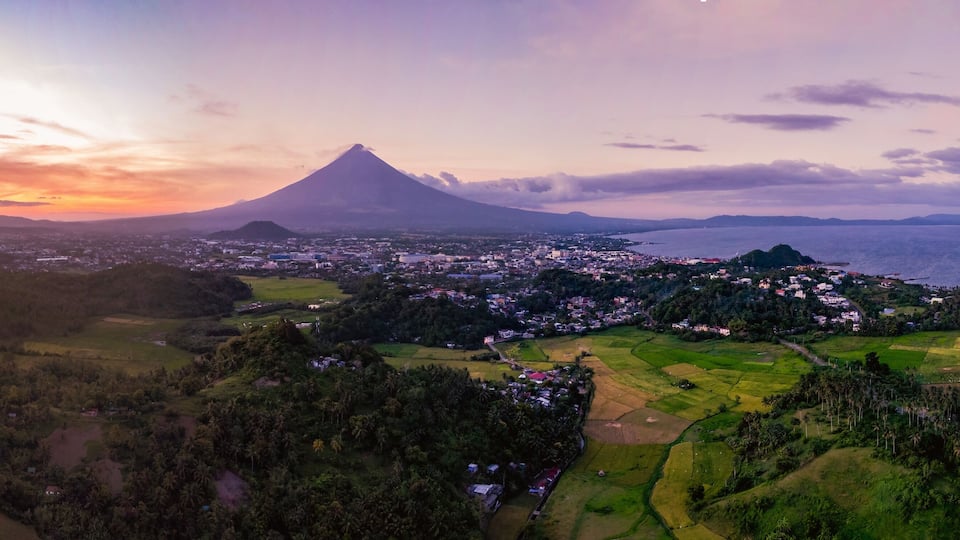 A volcano with a town in the foreground and a body of water in the background.