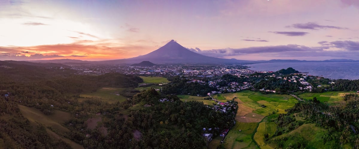 A volcano with a town in the foreground and a body of water in the background.