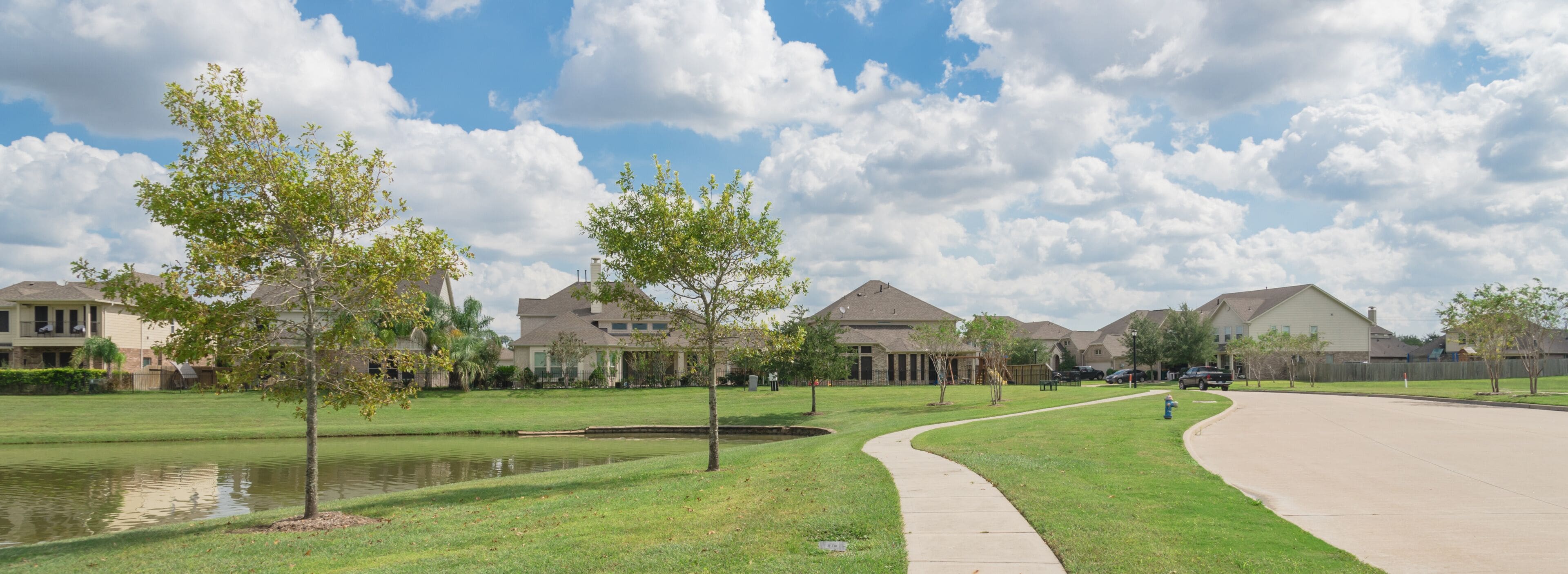 A suburban neighborhood with houses, a curved sidewalk, a small pond, and trees.