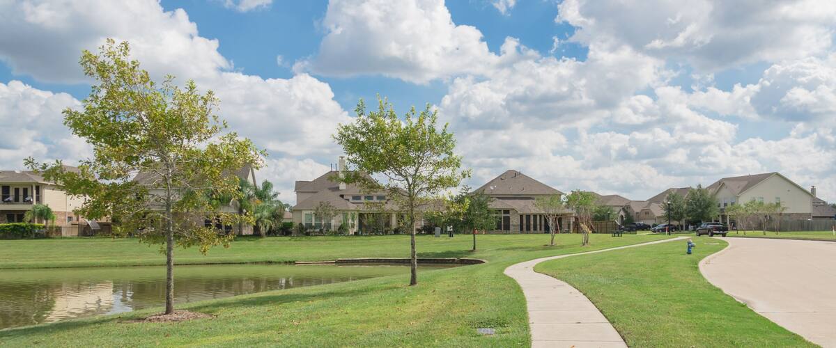 A suburban neighborhood with houses, a curved sidewalk, a small pond, and trees.
