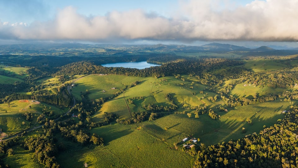 A lush green valley with rolling hills, a lake, and distant mountains under a partly cloudy sky.