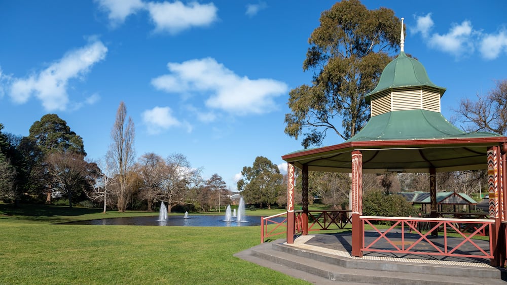 Ein Kiosk mit grüner Kuppel und roten Geländern in einem Park mit Springbrunnen und Bäumen.