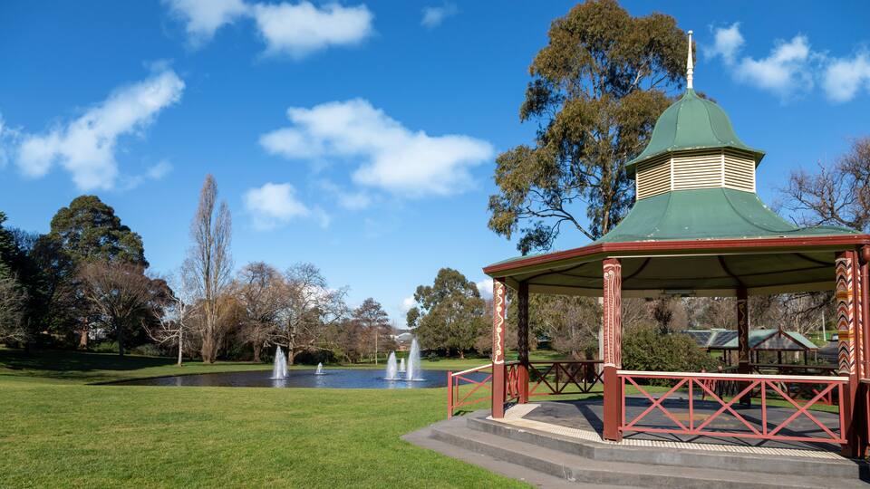 A gazebo with a green roof and red railings in a park with fountains and trees.