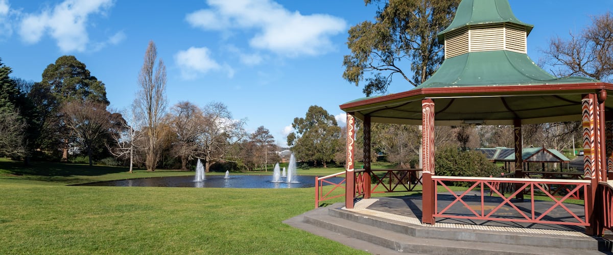 A gazebo with a green roof and red railings in a park with fountains and trees.