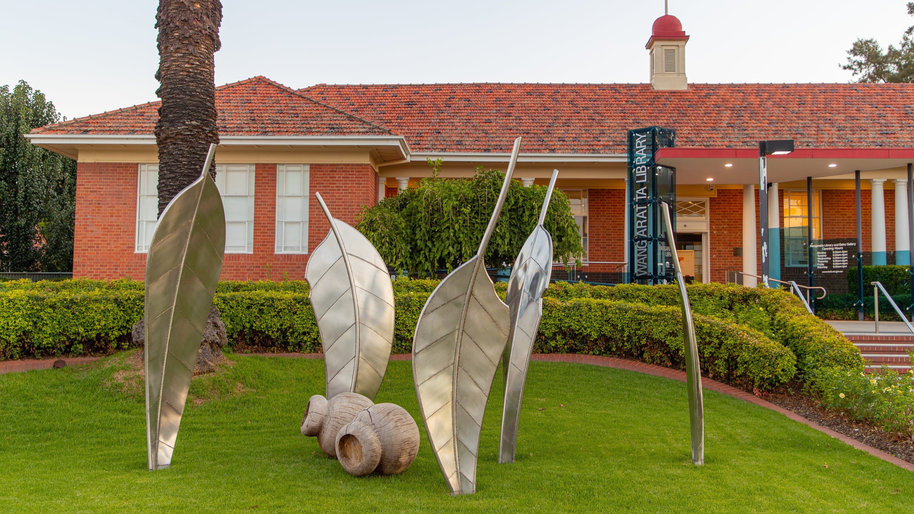 A building with a red-tiled roof, a sign reading "Wangara Library and Gallery," and a sculpture of abstract leaves in front.