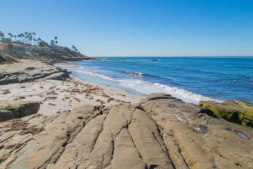 Una playa rocosa con olas, palmeras y casas al fondo.