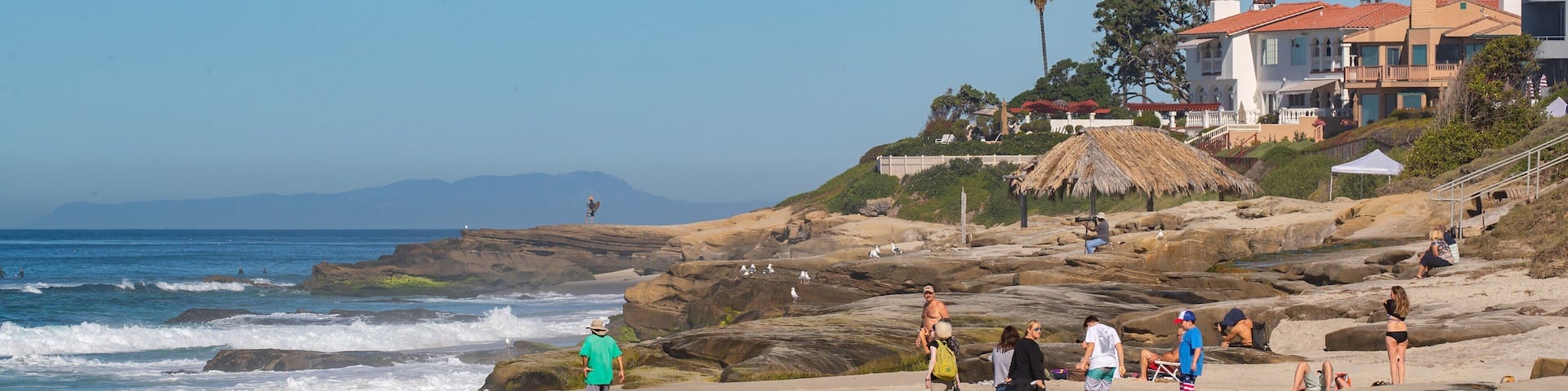 A rocky beach with people, waves, and a building with a thatched roof.