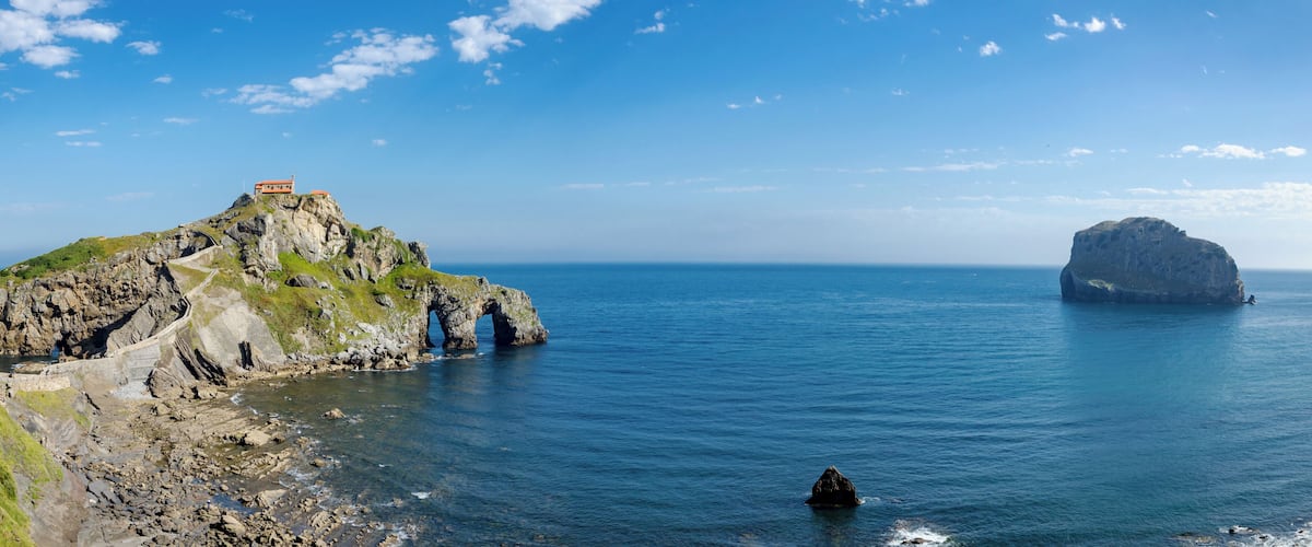 A rocky coastline with a lighthouse on a hill, a large rock formation in the sea, and a small boat in the water.