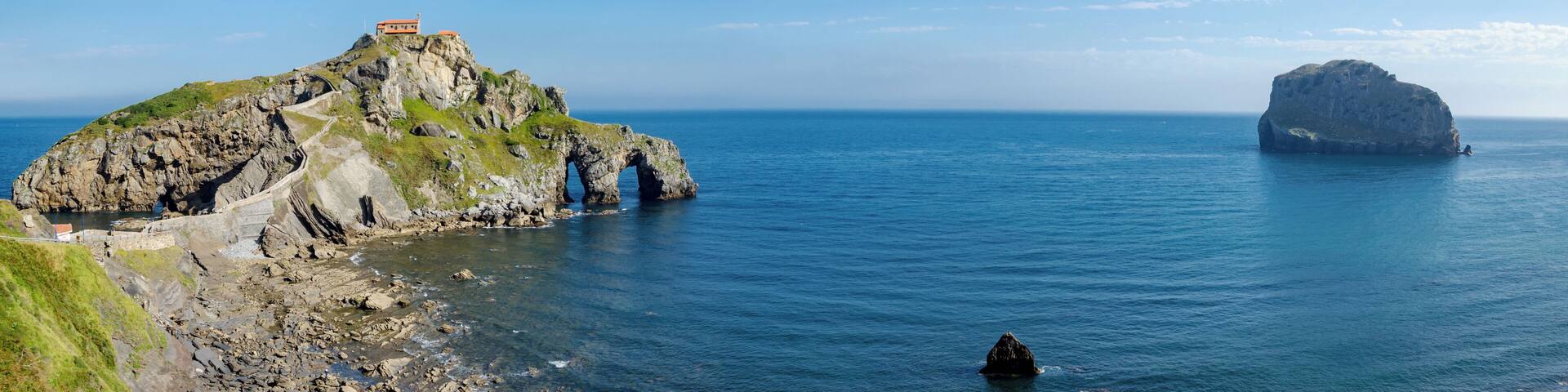 A rocky coastline with a lighthouse on a hill, a large rock formation in the sea, and a small boat in the water.