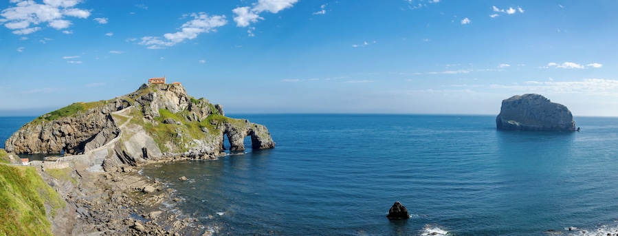 A rocky coastline with a lighthouse on a hill, a large rock formation in the sea, and a small boat in the water.