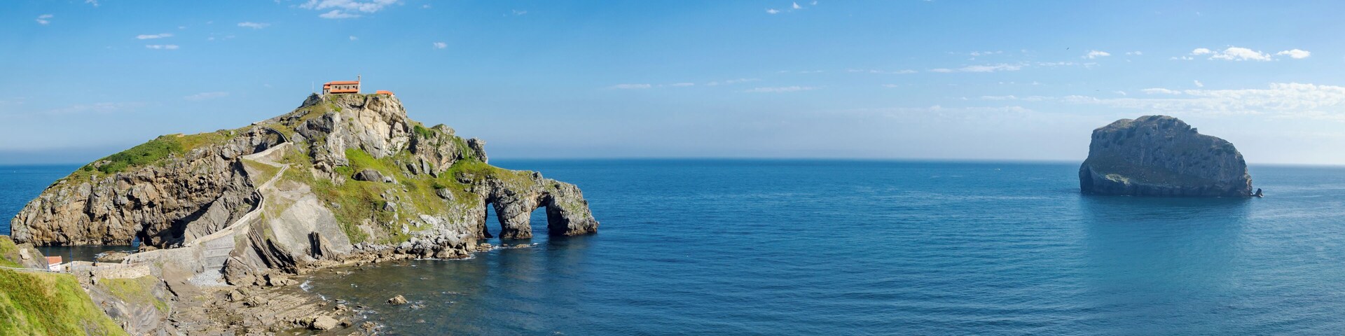 Costa rocosa con un faro en una colina, una gran formación rocosa en el mar y una pequeña embarcación en el agua.