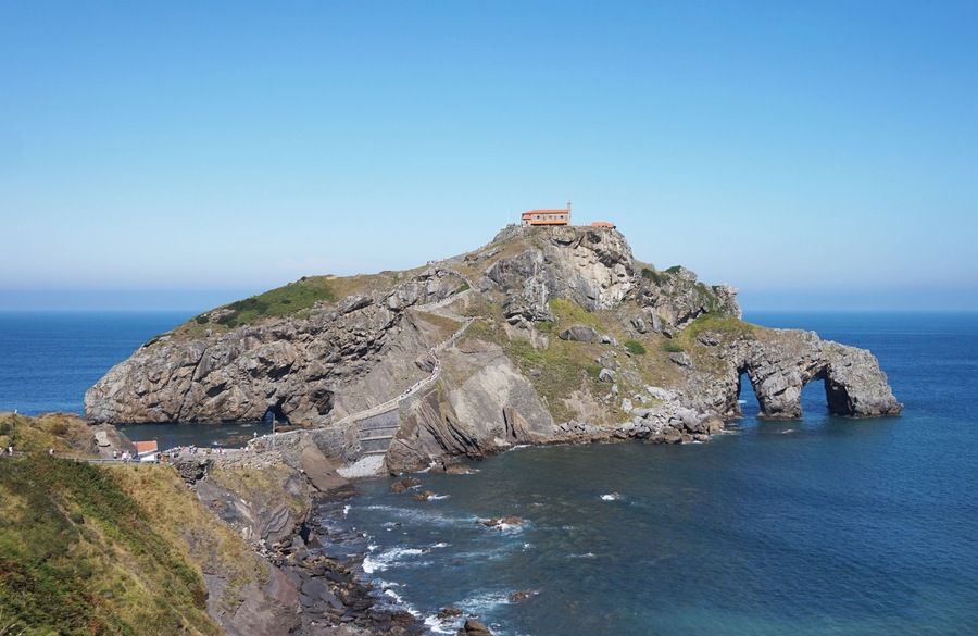 Una isla rocosa con un pequeño edificio en la cima, un arco natural en el mar y un camino costero.