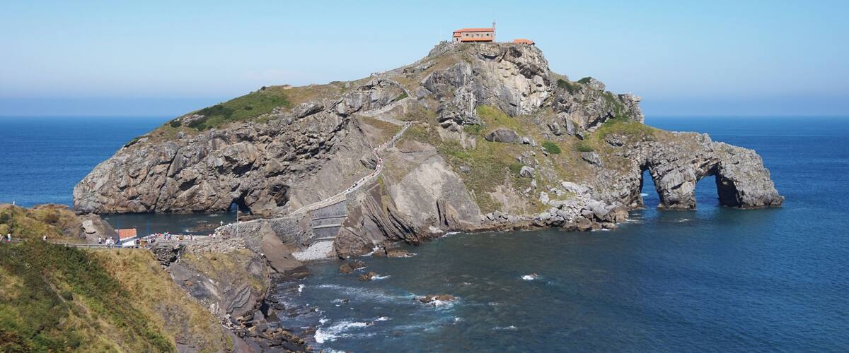 A rocky island with a small building on top, a sea arch, and a coastal path.