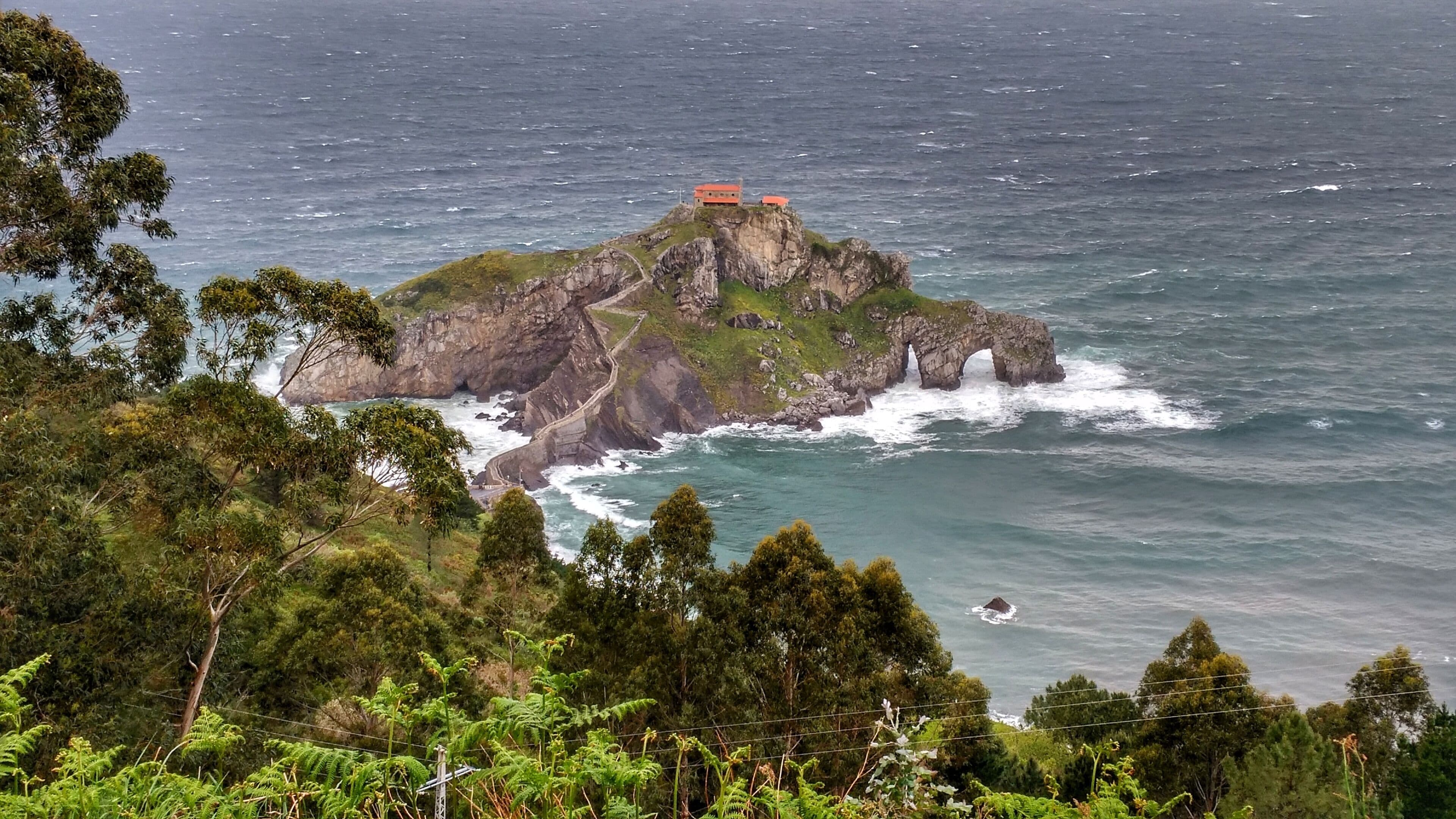 A rocky island with a small building in the distance, surrounded by ocean waves and dense greenery.