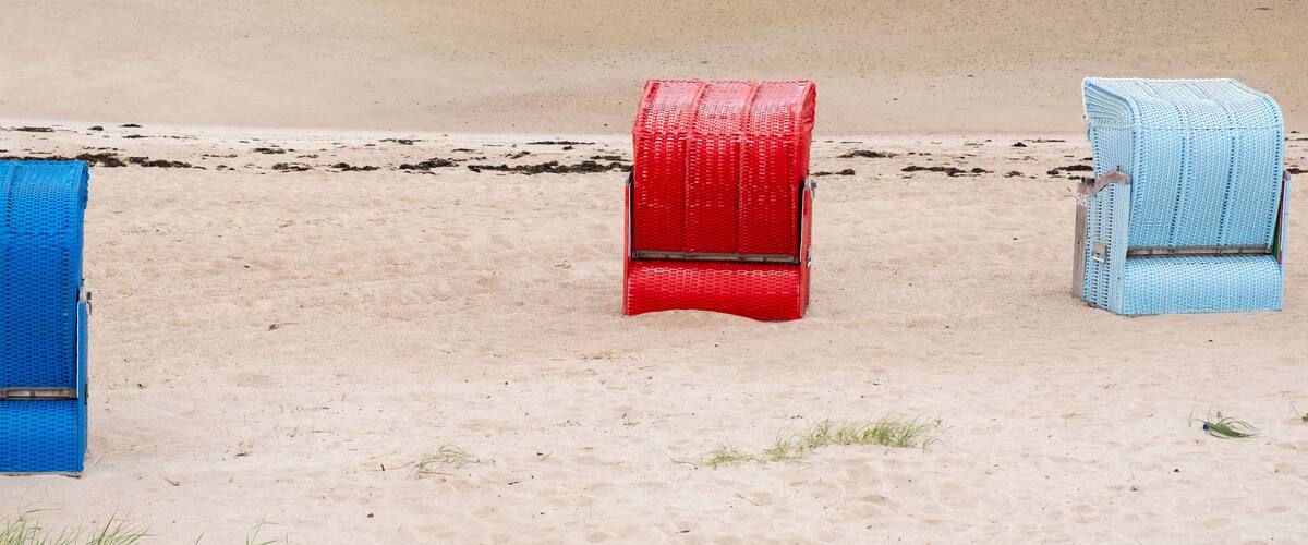 Three beach chairs in blue, red, and light blue colors on a sandy beach.