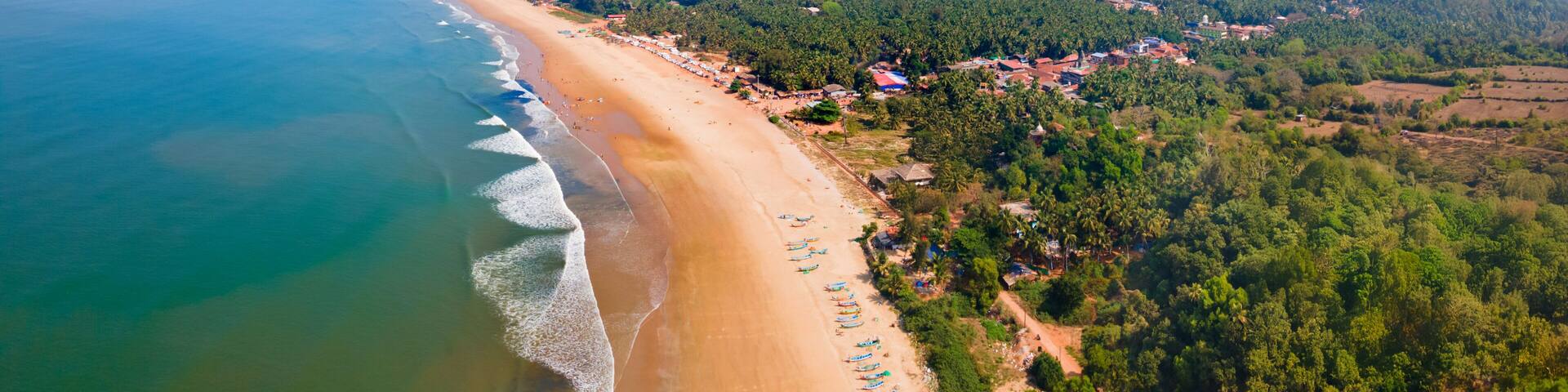 A coastal landscape with a sandy beach, a clear blue sky, and a backdrop of green hills and scattered buildings.