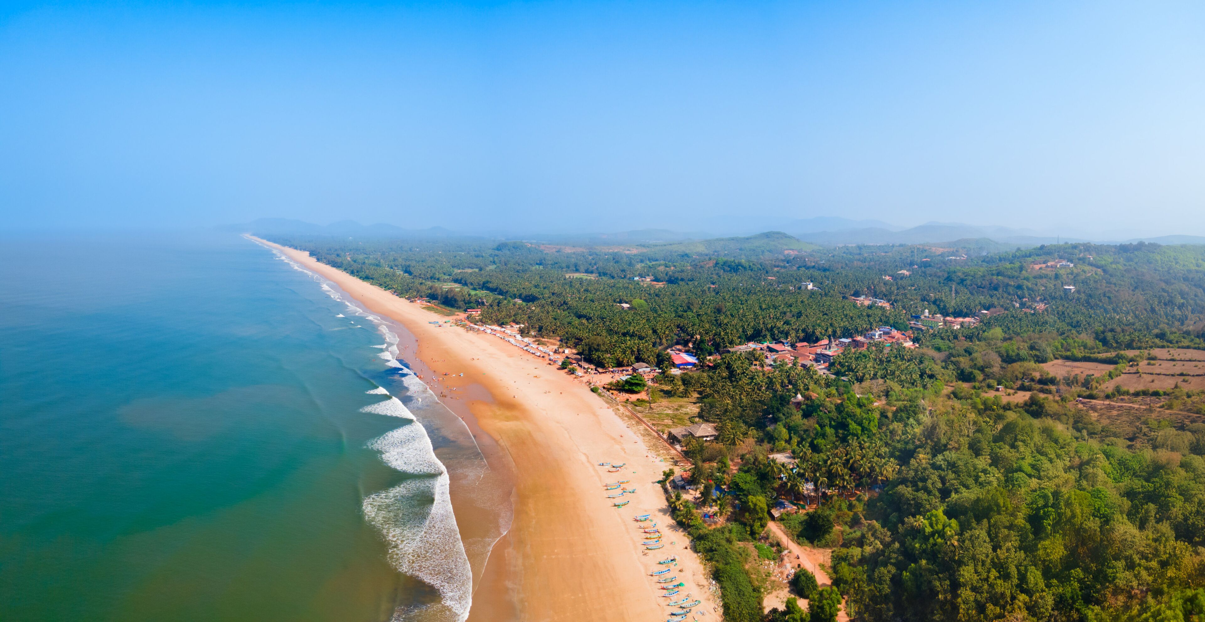 A coastal landscape with a sandy beach, a clear blue sky, and a backdrop of green hills and scattered buildings.