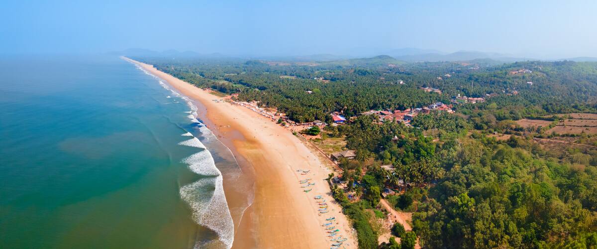 A coastal landscape with a sandy beach, a clear blue sky, and a backdrop of green hills and scattered buildings.