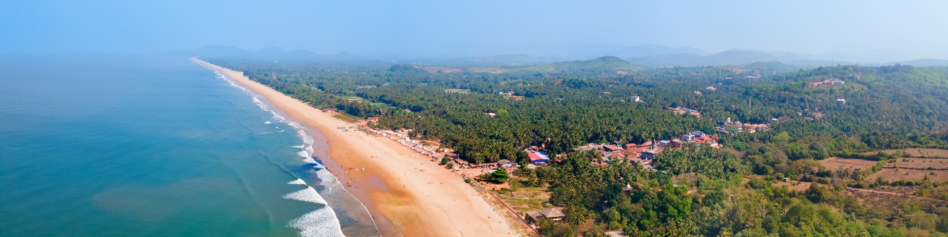 A coastal landscape with a sandy beach, a clear blue sky, and a backdrop of green hills and scattered buildings.