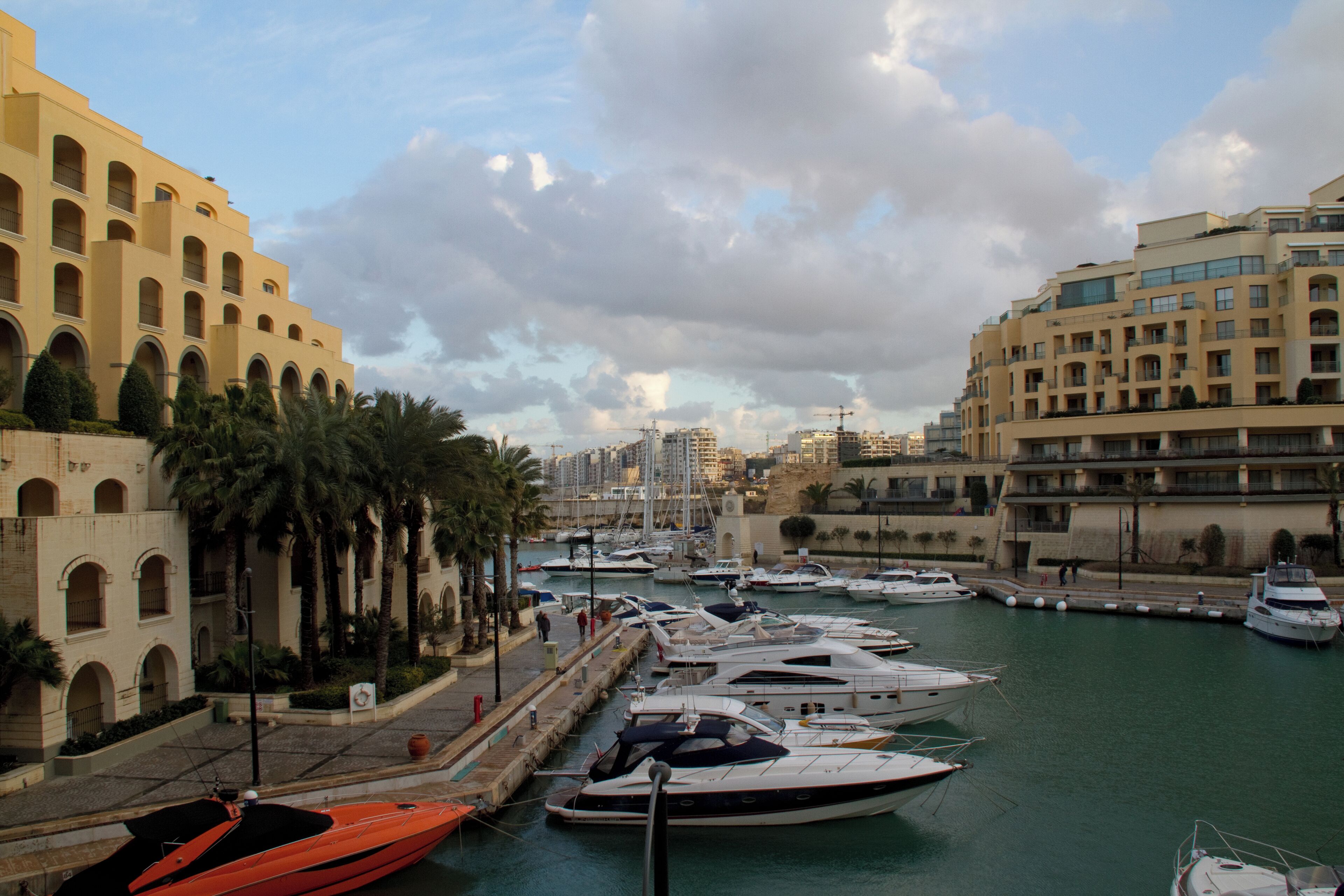 A marina with yachts docked, palm trees, and Mediterranean-style buildings.