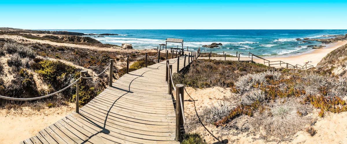 A wooden boardwalk leading to a beach with rocky coastline and sparse vegetation.