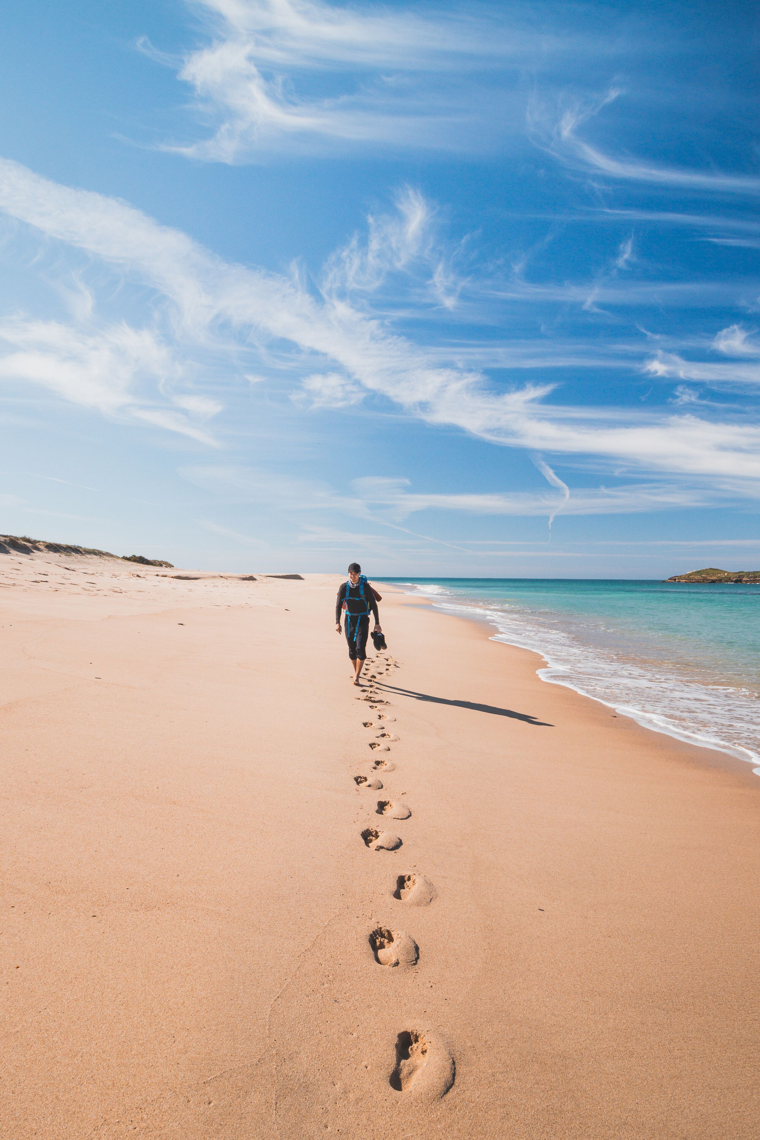 Passionate backpacker, backpack and boots in hand, walks along the Praia da Ilha do Pessegueiro beach on the Atlantic Ocean near Porto Covo, Portugal. In the footsteps of Rota Vicentina