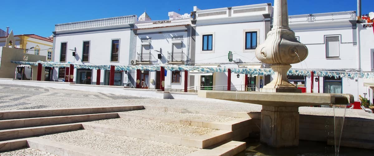A fountain in a plaza with steps leading up to it, surrounded by white buildings with balconies and windows.