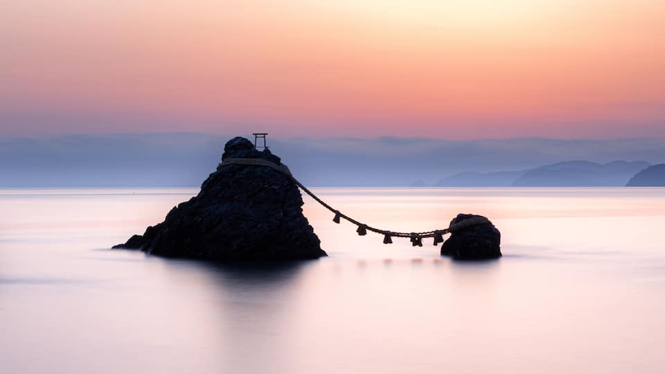 A rock formation with a chair and a rope bridge over calm water at sunset.