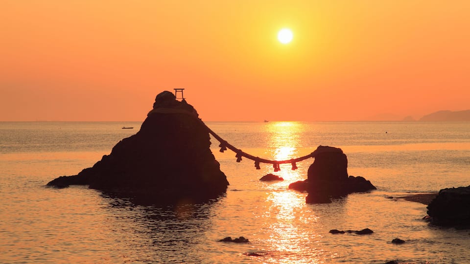 A sunset over a calm sea with rocky formations and a rope bridge.