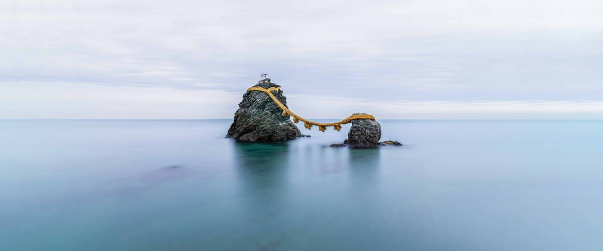 Two rock formations connected by a rope bridge over calm water.