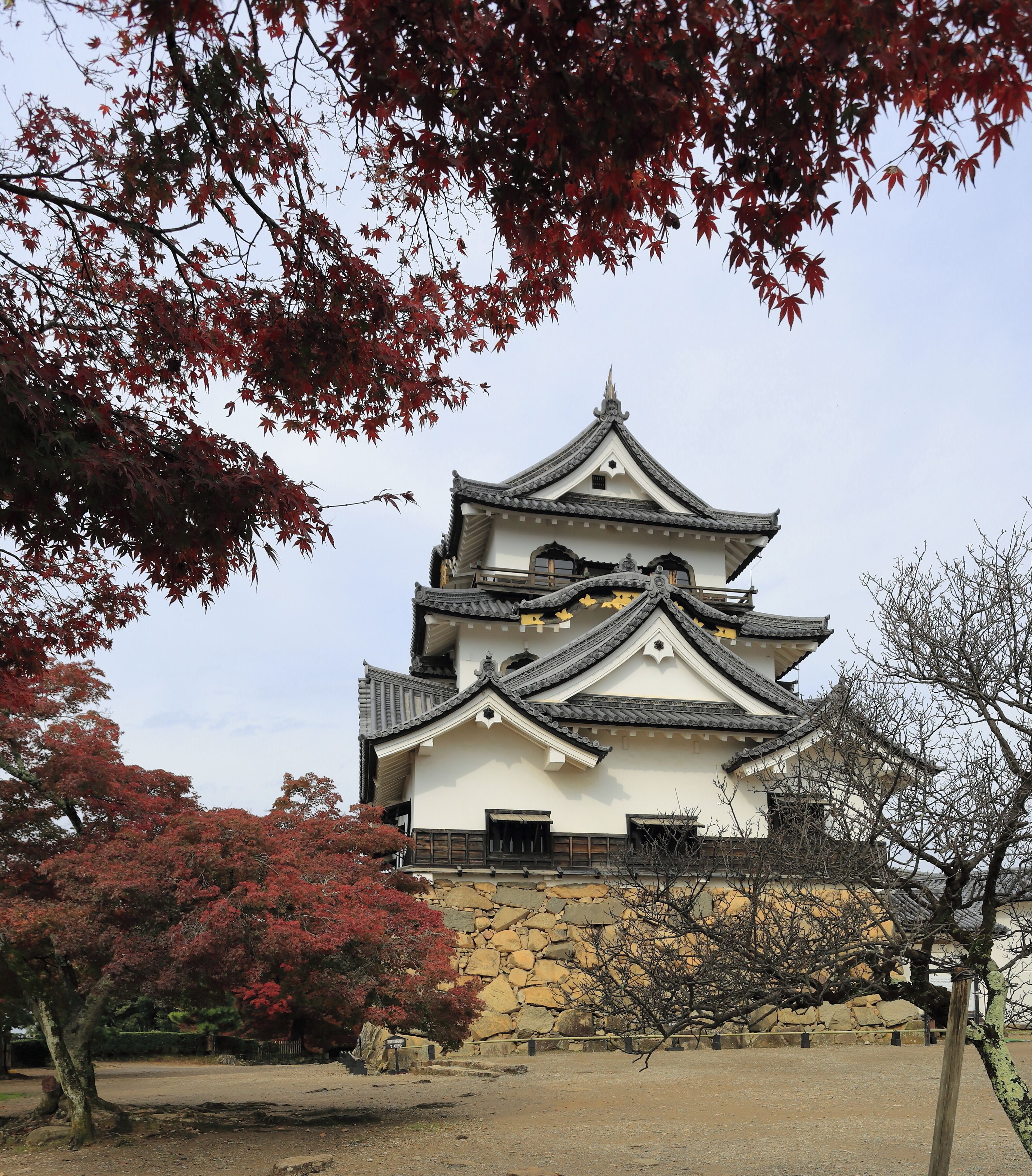 A traditional Japanese castle with multiple tiers and a stone base, surrounded by autumn foliage.