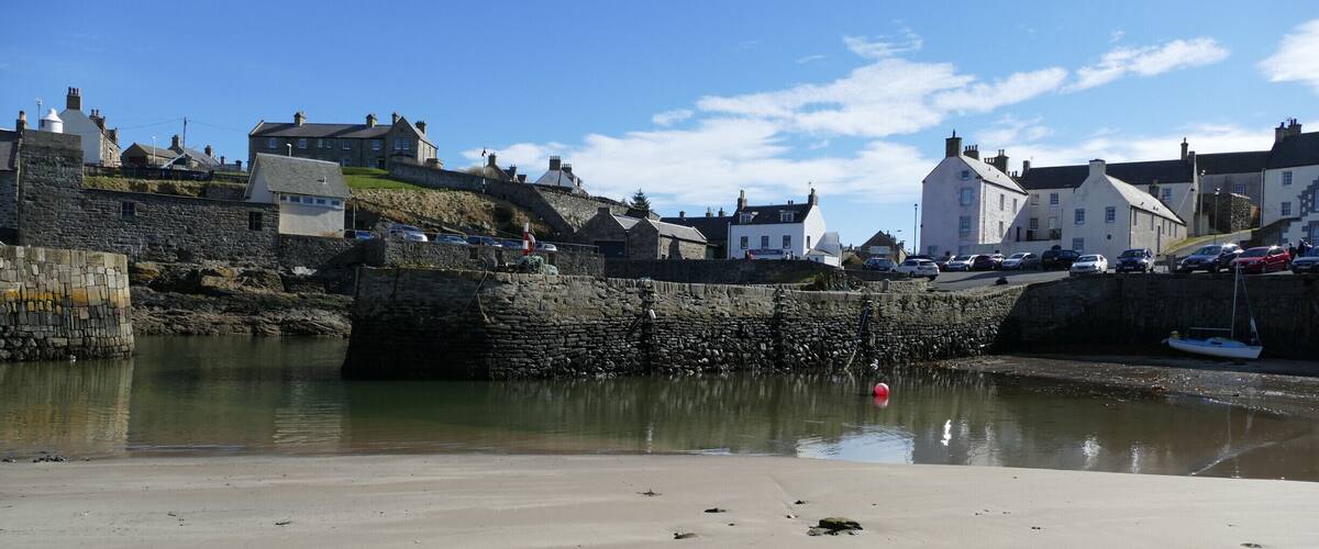 A sandy beach with footprints, a stone wall, and houses along the water's edge.