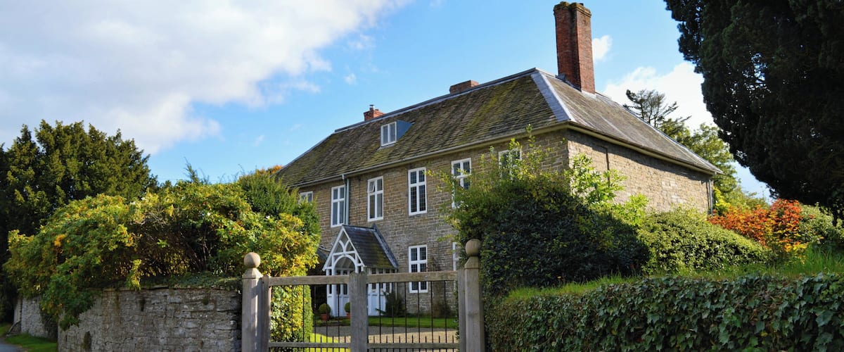 A stone house with a wooden gate, surrounded by lush greenery and a clear blue sky.